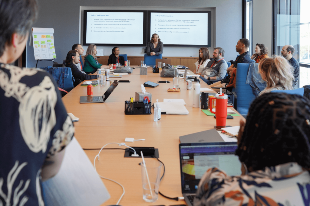 A group of people seated around a large conference table in a modern meeting room. A presenter stands at the front.